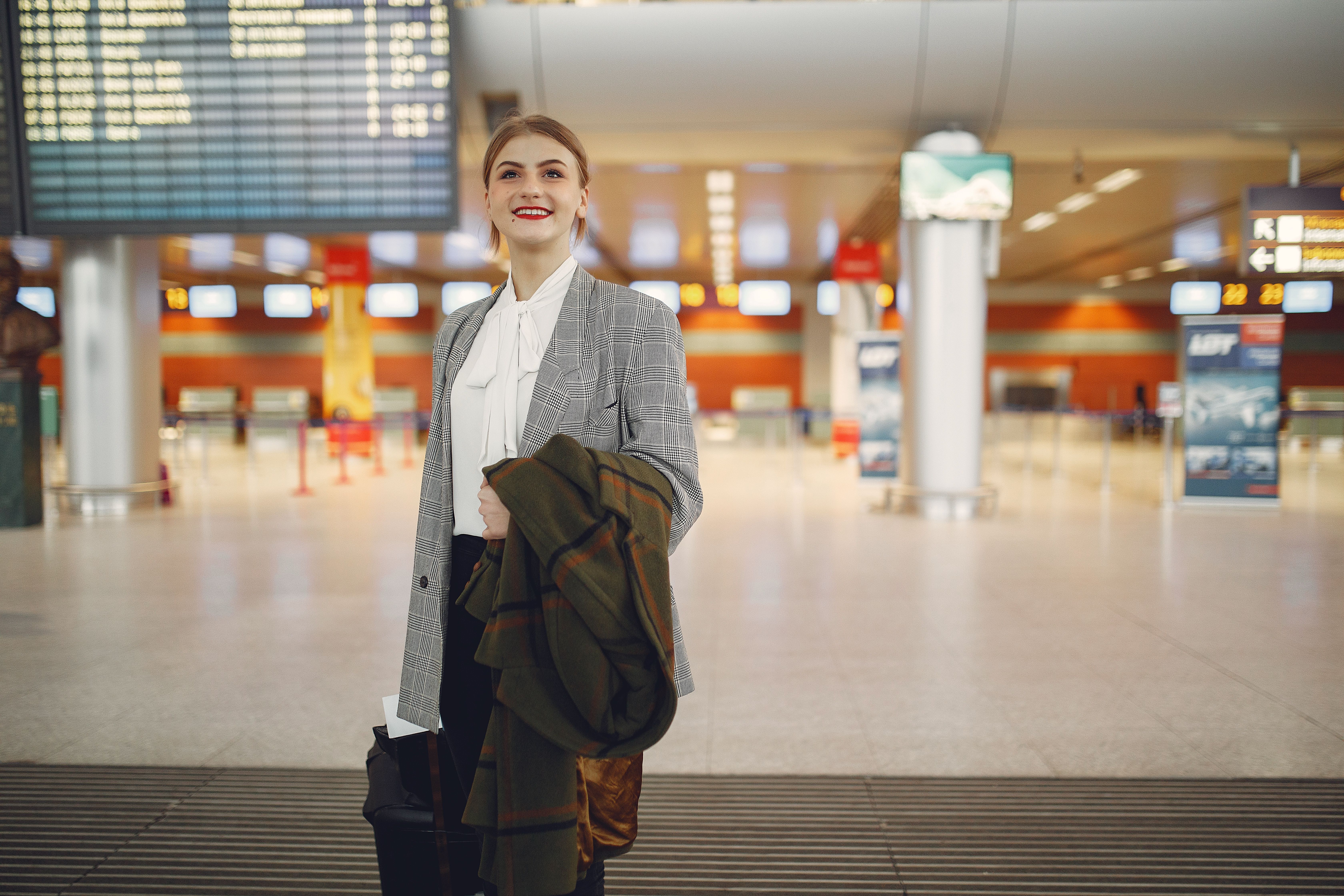 Smiling female manager checking flight schedule in an airport while standing with a suitcase and coat.